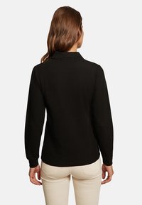 Černé polo tričko s dlouhým rukávem a límcem, hladká textura a přiléhavý design, nošené přes béžové kalhoty. Pohled zezadu ukazuje jednoduchost a eleganci.