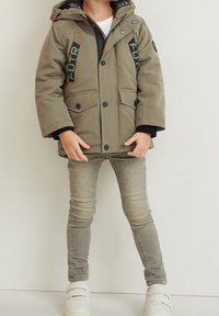 Khaki parka jacket with a hood, featuring large front pockets, black accents, and visible branding. Worn over a white shirt and gray jeans.