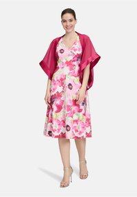 Blumenkleid mit rosa, roten und grünen Mustern; V-Ausschnitt und schwingendem Rock. Überlagert mit einem transparenten magentafarbenen Schal. Silberne Absätze sind sichtbar.