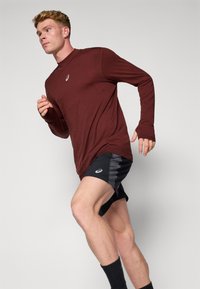 Dug dugih rukava boje bordo sportska majica u kombinaciji s crnim šortsevima s sivim detaljima. Model je u trkaćem položaju na jednostavnoj pozadini.