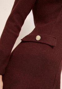Bordo pleteni haljina s teksturiranim materijalom, uskim siluetom i gumbom s bočne strane. Ima mali rever na izrezu.