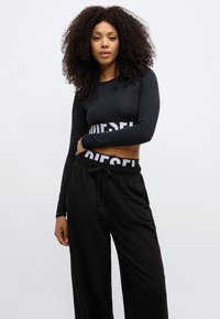 Melns cropped top ar garām piedurknēm un baltu "DIESEL" logotipu uz lentas, kombinēts ar vaļīgām melnām biksēm ar elastīgu jostasvietu.
