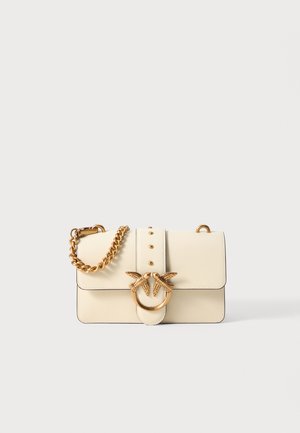 Cream leather handbag with gold chain strap, featuring a circular hardware accent with two bird figures and decorative studs on the front.