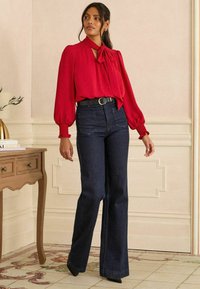 Crvena bluzica s detaljem mašne i napufanim rukavima u kombinaciji s tamnoplavi trapericama s visokim strukom i tregersima. Crni remen i cipele s oštrim vrhom.