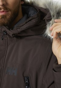 Hnědá zateplená bunda s kapucí s kožešinovým lemem, vysokým límcem, předním zipem a boční kapsou. Materiál vypadá robustně a voděodolně.