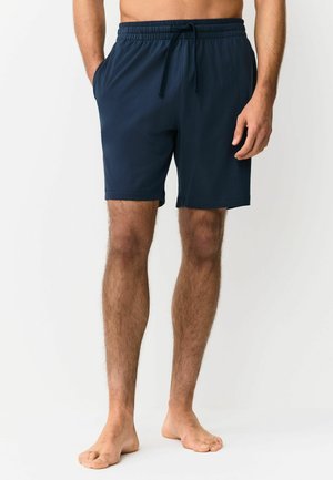 Bare-chested person standing barefoot wearing navy blue drawstring shorts with hands in pockets against a plain white background.