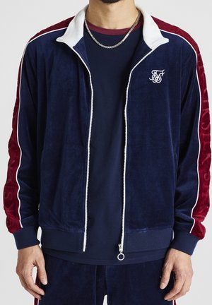 Veste de survêtement velours bleu marine avec des accents en velours rouge, zip blanc et logo brodé sur la poitrine gauche. Elle dispose d'un col montant et de poignets côtelés.