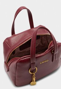 Sac à main en cuir bordeaux avec deux poignées courtes, une fermeture éclair dorée et un petit charm. L'intérieur présente une doublure à motif logo.