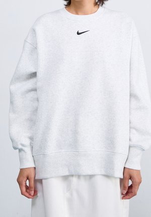 Osoba nosząca jasnoszarą bluzę Nike z małym czarnym logo Swoosh umieszczonym na środku przy dekolcie, w połączeniu z białymi spodniami.