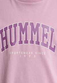 Lyserød bomulds t-shirt med fremtrædende lilla tekst "HUMMEL" og sætningen "SPORTSWEAR SINCE 1923" i mindre hvid skrift nedenfor.