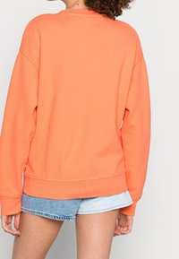 Mujer vistiendo un sudadera de gran tamaño en un brillante color naranja y un pantalón corto de mezclilla en dos tonos, vista desde atrás contra un fondo liso.