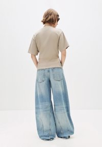 Person med kort hår iført solbriller, beige t-shirt og lyseblå vide jeans står med hænderne i baglommerne mod en ensfarvet baggrund.