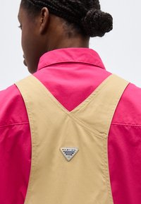 Άτομο με πλεγμένα μαλλιά που φοράει μπεζ ολόσωμη φόρμα πάνω από ένα φωτεινό ροζ πουκάμισο με γιακά, από πίσω, με λογότυπο σε σχήμα τριγώνου.