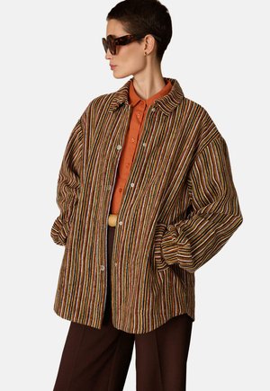 Femme aux cheveux courts portant de grandes lunettes de soleil en écaille, une veste rayée oversize, une chemise orange rouille à boutons et un pantalon marron foncé.