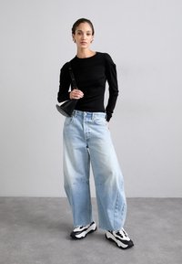 Svart langermet topp, lyseblå vide jeans, svarte og hvite joggesko og en svart håndveske, stående foran en grå bakgrunn.