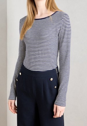 Vrouw met een marineblauw-wit gestreepte longsleeve en marineblauwe hoge taille broek met decoratieve gouden knopen.