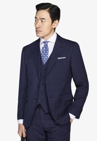 Mężczyzna w granatowym trzyczęściowym garniturze z niebieskim krawatem w wzory i białą poszetką, trzymający czarną torbę, patrzący w lewo.