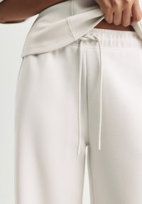 Persona che sistema pantaloni sportivi beige chiaro con coulisse in vita, indossando una maglietta abbinata, mostrata dalla zona addominale alle cosce su sfondo bianco.