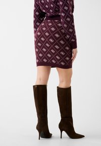 Pletená mini sukně v barvě bordó s diamantovým vzorem, doplněná o hnědé semišové boty na podpatku po kolena se zapínáním na zip na straně a zlatými detaily.