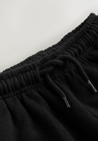 Črne športne hlače imajo teksturiran, elastičen pas z vrvico in kovinskimi konicami, kar poudarja mehak in udoben material.