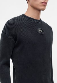 Homme portant un pull noir côtelé avec un petit emblème ovale en métal et tissu sur la partie supérieure de la poitrine, sur un fond clair uni.