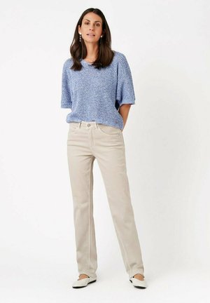 Woman standing wearing light blue short-sleeve knit top, beige straight-leg pants, and white flats against a white background.