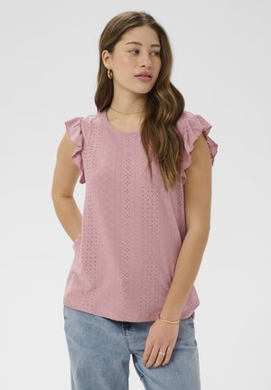 Fiatal nő hosszú barna hajjal, rózsaszín áttört ujjatlan felsőt visel fodros vállakkal és világoskék farmert, oldalra néz.