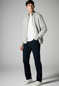 Graue Zip-Up-Jacke mit hohem Kragen, getragen über einem weißen T-Shirt, kombiniert mit dunkelblauen Jeans und weißen Sneakern. Glatte Textur.