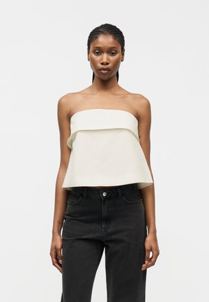 Femme portant un haut blanc sans bretelles avec un détail plissé, associé à un jean noir taille haute, debout devant un fond clair uni.