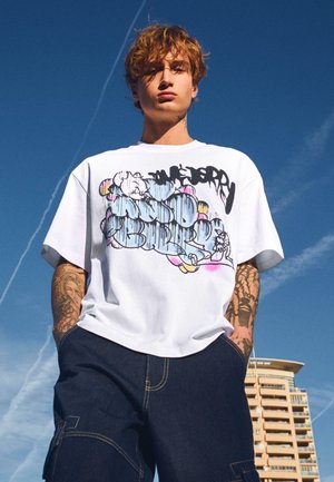 Mladý muž s tetovaniami oblečený v bielom tričku s potlačou a tmavých nohaviciach stojí pred jasne modrou oblohou a vysokou budovou.