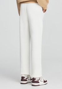 Białe dresowe spodnie o luźnym kroju, z elastycznymi mankietami i kieszeniami bocznymi, zestawione z brązowo-białymi sneakersami. Miękka, gładka tekstura materiału.