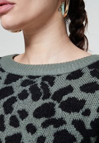 Stickad tröja i olivgrön med svart leopardmönster, med en ribbad halsringning. Mjuk textur med en avslappnad passform och korta ärmar.