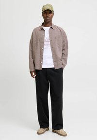 Chemise à carreaux marron sur un t-shirt graphique blanc, associée à un pantalon noir et des chaussures beiges, le tout couronné par une casquette verte.