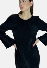 Vestido negro con una tela suave y texturizada; escote redondo y mangas de campana, con un corte relajado que llega hasta la mitad del muslo.