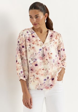 Frau mit dunklen Haaren im Pferdeschwanz, trägt eine geblümte V-Ausschnitt-Bluse mit Puffärmeln und weiße Hose, Hände entspannt, eine in der Tasche.