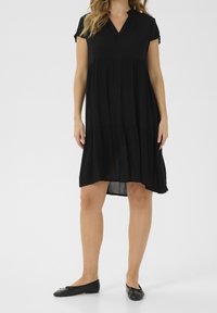 Robe noire à manches courtes avec un décolleté en V, présentant un design en étages. Fabriquée en tissu léger, elle a un ourlet arrière légèrement plus long. Assortie à des ballerines noires.