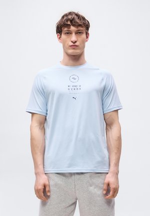 Jeune homme portant un T-shirt bleu clair avec l'inscription "My Sport is HYROX" et un short gris, debout devant un fond clair uni.
