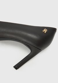 Must musta kõrgepaar nahast kingad terava nina, kuldse logoga detail ja sile, sujuv tekstuur. Toode on varustatud peenikese kontsaga, et anda elegantsi.