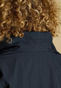 Jachetă ușoară de marină cu guler înalt, având „JOULES” brodat în negru pe spate. Textură netedă cu detalii minime.