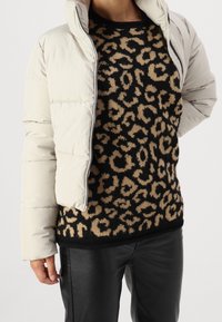 Veste crème matelassée avec un col montant, portée sur un pull noir orné d'un motif léopard beige et un pantalon en similicuir noir.