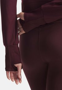 Burgunderfarbenes Langarmoberteil mit gerippten Bündchen, kombiniert mit glatten Leggings. Der Stoff ist dehnbar und enganliegend, wodurch die Konturen betont werden.