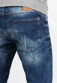 Jean en denim de lavage foncé avec des sections décolorées, présentant un design de poche arrière cousue et une étiquette en cuir brun clair au niveau de la taille.