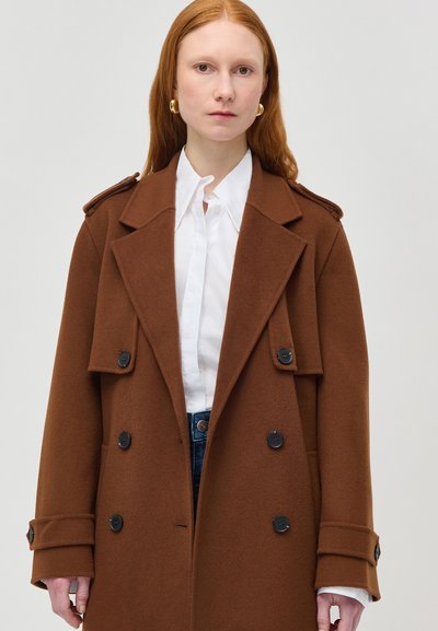 Donna con capelli rossi lunghi che indossa orecchini a cerchio d'oro, una camicia bianca e un cappotto di lana marrone doppiopetto con bottoni neri.