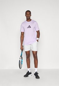 Jasnofioletowa koszulka Adidas z czarnym logo, zestawiona z szarymi szortami. Model trzyma rakietę tenisową i ma na sobie czarne buty oraz białe skarpetki.