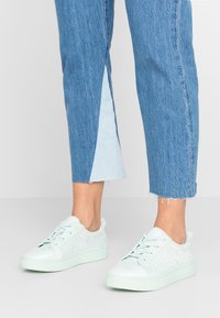 Jasno miętowe sneakersy z zamszową fakturą, z niską cholewką i okrągłym noskiem. Zestawione z niebieskimi, ściętymi spodniami dżinsowymi.