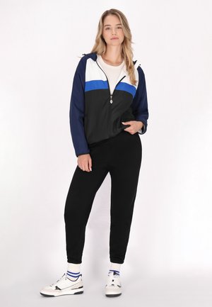 Vindjakke med hvide, blå og sorte farveblokke; lynlåslukning; justerbar hætte; parret med sorte joggers og sneakers.