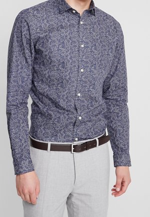 Mand iført en marineblå langærmet skjorte med et lille hvidt blomsterprint, lysegrå bukser og mørkebrun bælte med sølvspænde.