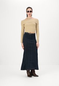 Beige avkortet genser med off-shoulder design, kombinert med en mørk denim maxiskjørt med vertikale sømdetaljer og en metallspenne. Brune ankelstøvler.