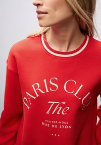Rode sweatshirt met witte letters "PARIS CLUB" en "The" in een opvallend lettertype. Bevat een geribbelde halslijn met contrasterende strepen.