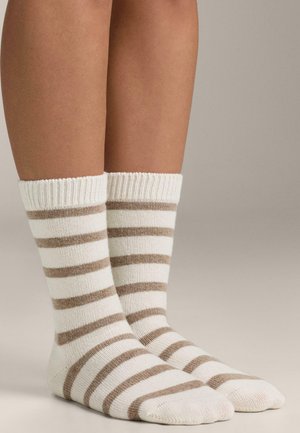 Beine mit kniehohen gestrickten Socken mit horizontalen beige-weißen Streifen vor einem neutralen Hintergrund.